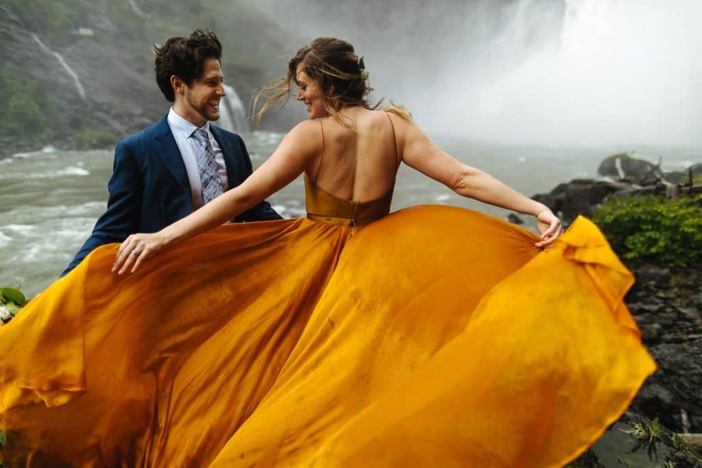 Groom and bride laugh while orange wedding dress is whipped in waterfall during washington state elopement